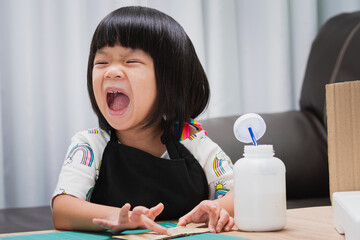 Happy Asian girl wearing black apron, child laughter while gluing on a piece of craft. The concept of learning through play. Kid aged 4-5 years old.