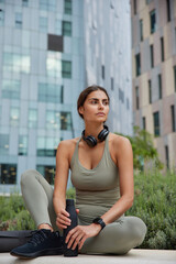 Fototapeta premium Pensive young woman gets ready for running routine holds bottle of fresh water feels thirsty after cardio training dressed in activewear poses against cityscrapers outdoor. Sport activity in open air