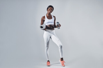 Fototapeta na wymiar Happy young African woman in sports clothing holding bottle with water and exercise mat