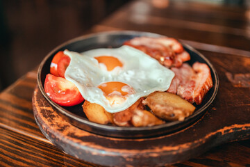 Fried eggs with bacon, tomatoes and toasts on plate on wooden table in a cafe. English breakfast