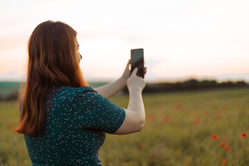 Back view of a young girl in a summer dress makes a photo on her smartphone in an autumn field at the golden hour