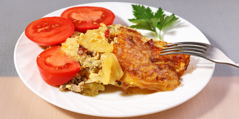 Portion of potato casserole with meat, golden crust on plate with fresh tomatoes and greens