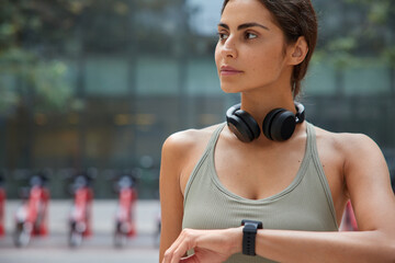 Cropped shot of sporty woman checks workout on smartwatch uses headphones to listen music from playlist waits for coach to start training monitors pulse after jogging exercises wears sportswear