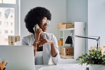 Beautiful young African woman talking on mobile phone and smiling while sitting at her working...