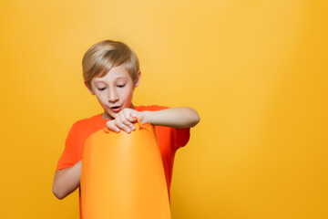 Obraz premium a boy in an orange T-shirt and on a yellow background holds a shopping bag in his hands looks into it with surprise