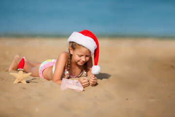 The child sits on the sand wearing a Santa hat. Little girl smiles and looks at a box with a gift. Merry Christmas. Winter vacation. Sea in the background. Copy space.
