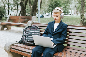 Fototapeta premium cute schoolboy with backpack in a school uniform with headphones and laptop is sitting on bench in the school yard, concept back to school and start of lessons