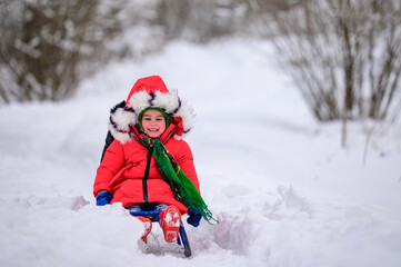 Little girl sitting on a sleigh on a snowy winter day, warm winter clothes, a red jacket and a green scarf.