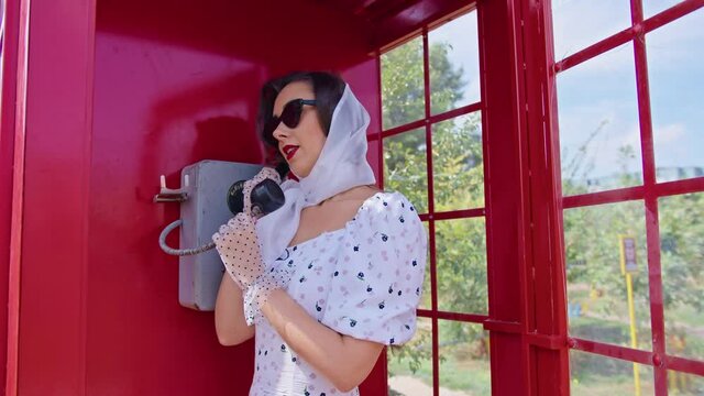 beautiful young woman happily speaks on the phone in an english style red telephone booth. girl dressed in a white dress and glasses. pinup.