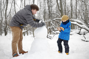 Fototapeta na wymiar Little boy with his father building snowman in snowy park. Active outdoors leisure with children in winter. Kid during stroll in a snowy winter park