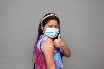 Happy Hispanic 10-year-old girl with mask shows her arm that just received the Covid-19 vaccine in...