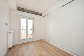 Bedroom with wall covered by a large white built-in wardrobe on one side and window leading to the...