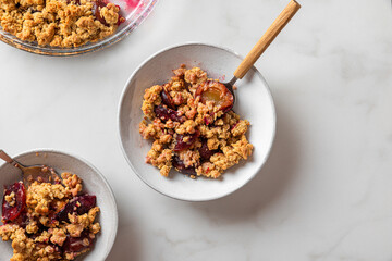 Fruit crumble. Plum crumble pie in plates with spoons on white marble table for tasty breakfast....