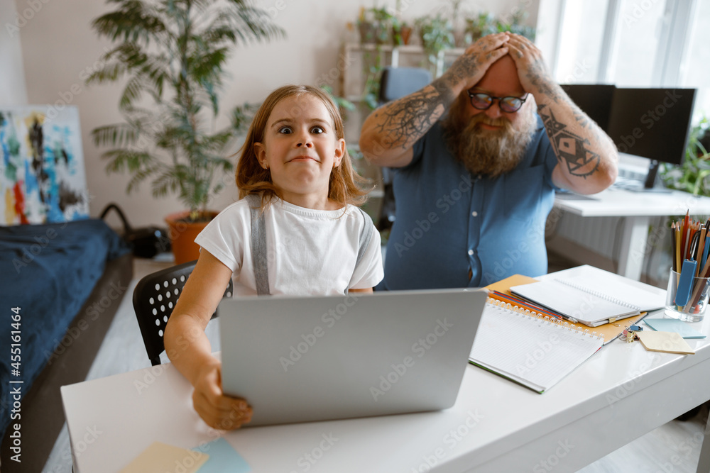 Wall mural Surprised little girl does homework on laptop while father clutches head at home