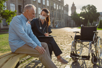 Young care taker nurse wearing face shield chatting with aged recovering male patient while sitting together on the bench in park near hospital