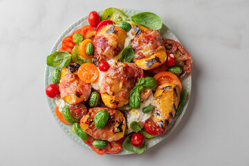 Grilled peaches and prosciutto salad with burrata cheese, tomatoes, cucumber and basil in a plate on white table. Top view