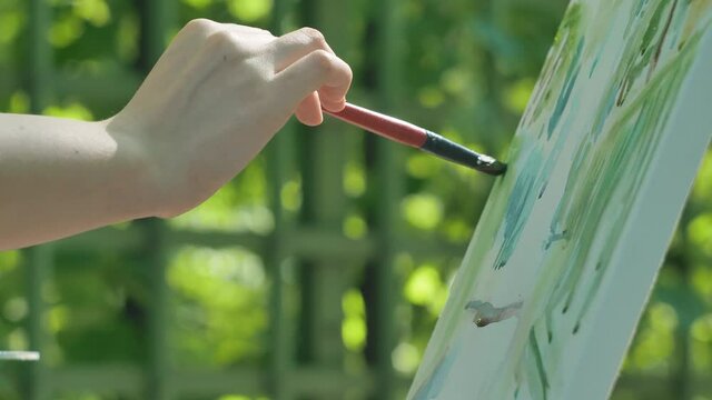Artist colours canvas on easel with special paintbrush in park against blurred green trees at back sunlight extreme closeup