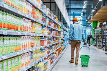 African-American guy with green plastic basket walks looking at shelves with different goods in...