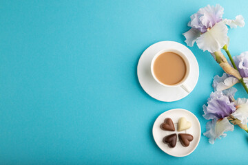 Obraz na płótnie Canvas Cup of cioffee with chocolate candies and lilac iris flowers on blue pastel background. top view, copy space.