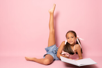 Beautiful little girl lying on the floor with one leg up studying isolated over pink background.