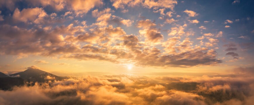 Mountains in low clouds at sunrise in summer. Aerial view of mountain peak in fog. Beautiful landscape with high rocks, forest, colorful sky. View from above of mountain valley in clouds. Foggy hills