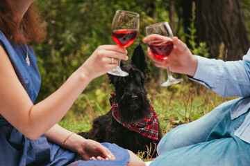 A couple in love and a dog on a picnic in the park. A man and a woman are holding glasses of red wine.