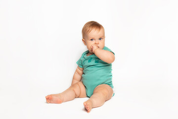 cute Caucasian one year old baby sucks thumb and scratches his first teeth. toddler boy teasing on white background in studio. baby care and child health concept