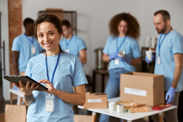 Portrait of happy young female volunteer in blue uniform using tablet pc and smiling at camera while standing indoors. Team sorting, packing items in the background