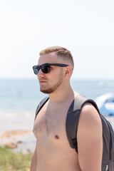  Young stylish man in a black sunglasses walking and relaxing on a beach. Relaxed and cheerful. Outdoor portrait of happy young caucasian man near seaside,  Stubble and blacks hair. Summertime.