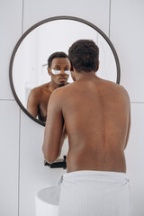 Attractive young African-American man with bare torso uses under eye patches looking in round mirror in bathroom close view
