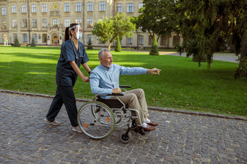 Full length shot of caring nurse wearing face shield and mask spending time with senior male patient in wheelchair on a walk in the park near hospital
