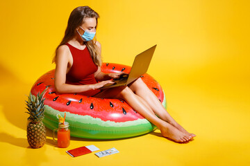  young woman dressed in red swimsuit sitting on inflatable circle on the yellow backgroun. wearing a mask with a laptop near the cocktail pineapple tickets looking at the laptop 