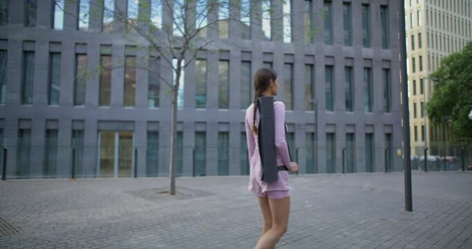 Slim active young woman walks around city has pilates exercisess with fitness mat passes by modern building walks to sports club alone covers long distance per day goes to location for workout