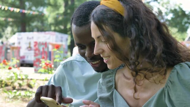 Black man and brunette young woman watch photos in smartphone and discuss laughing in attraction park on sunny day closeup