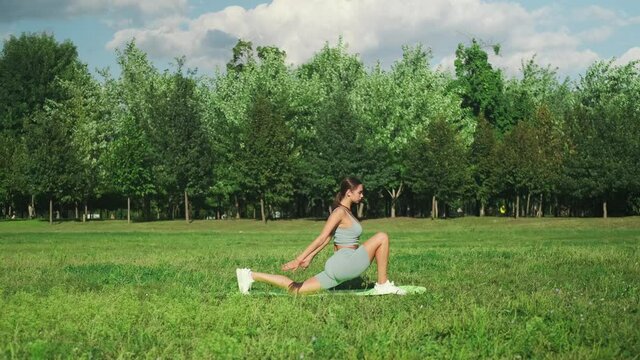 A beautiful sportive girl is engaged in yoga on green grass in a city park. Ashva sanchalanasana pose. Slow motion. 4k footage