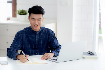 Young asian business man writing on notebook for planning working and using laptop computer on desk at home, notes about finance, male study and learning, business and communication concept.