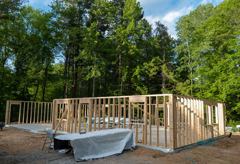 New home construction site with wood studs of outer walls in place, and contractors tools and materials on the concrete slab. Trees line the back of the building site..