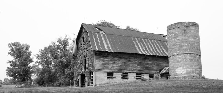 Black and white image of dreary old farm barn and silo with overcast sky in northern Minnesota