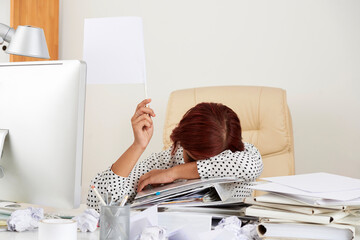 Exhausted young businesswoman sitting at table with face on stack of documents and showing white flag - Powered by Adobe