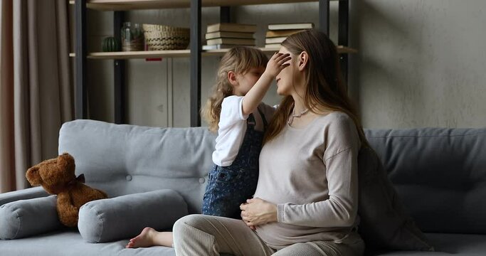 Young pregnant woman sitting on sofa with preschool 4s adorable daughter, kid touches big belly of mother talking hugging enjoy tender moments and free time together at home. Happy motherhood concept