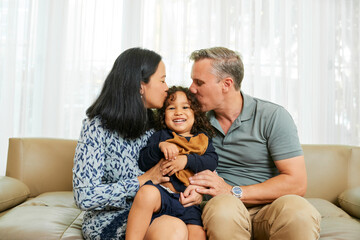 Mother and father kissing their adorable happy little son on both cheeks when sitting on sofa at home - obrazy, fototapety, plakaty