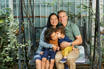 Happy family of mother, father and two little sons sitting on swing in backyard and smiling at...