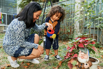 Smiling young mother teaching son how to water plants and flowers in backyard with watering can