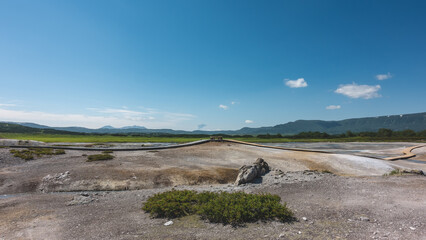 Fototapeta premium The caldera of an extinct volcano has rocky soil. Plank paths are laid for tourists. In the distance there is a green meadow. A mountain range against the blue sky. Kamchatka. Uzon