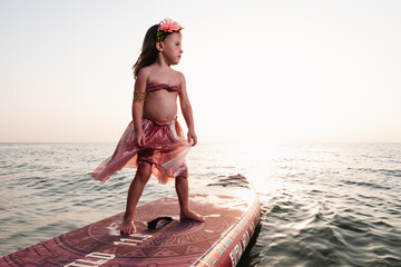 Little girl on a surfboard in the sea at sunset. In Moana's cast.