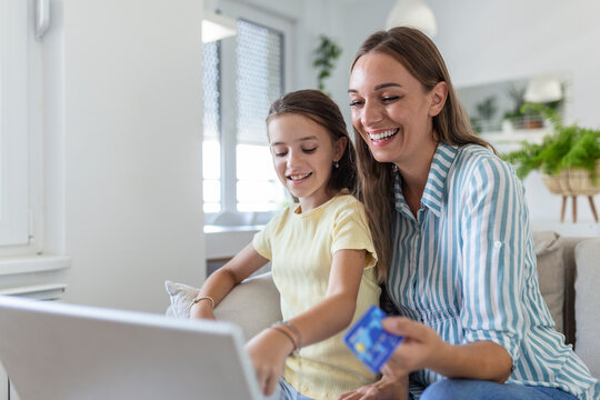 Low angle of happy small daughter rejoicing while smiling mother in casual clothes focusing on screen and using laptop for online shopping in light modern apartment