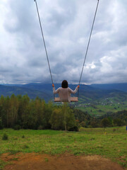Back of girl with dark hair in a gray sweater sitting on a swing with the mountain view. Calm and quiet wanderlust concept moment when person feels happiness and life. Alone travel in Ukraine.