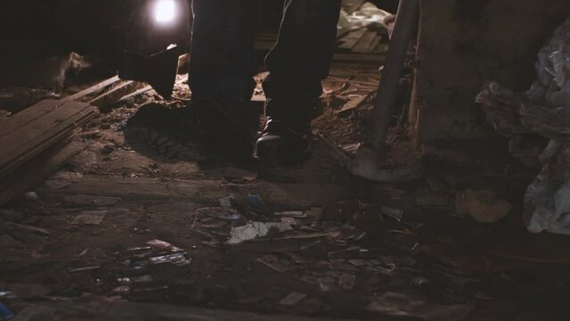 A man in heavy boots with an axe in his hand walks through a dusty, abandoned house. In the background are a pickaxe, trash, and cobwebs. The concept of a homicidal maniac