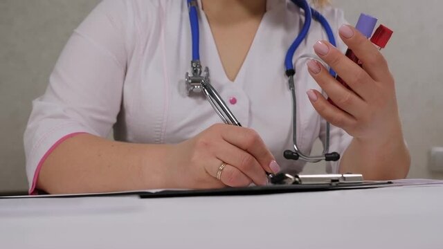 Close-up of doctor arms holding blood test tube and making notes. Woman is sitting at table in lab. Nurse writing the prescription after blood test.