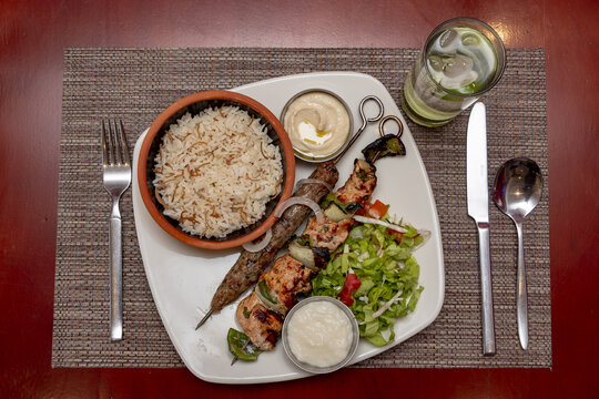 Top View Of Shish Taouk, Kebab, Rice, And Cutlery On A White Plate On A Red Table